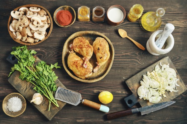 Top view of fried chicken thighs on a wooden plate. Chopped onions, mushrooms, and herbs are placed nearby. Various spices and oil are in jars ready for cooking