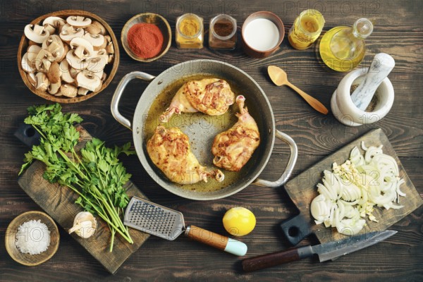 Roasted chicken legs in a pan on a wooden tabletop surrounded with mushrooms, onions, and various spices. Fresh herbs and a lemon are also on the table ready for use