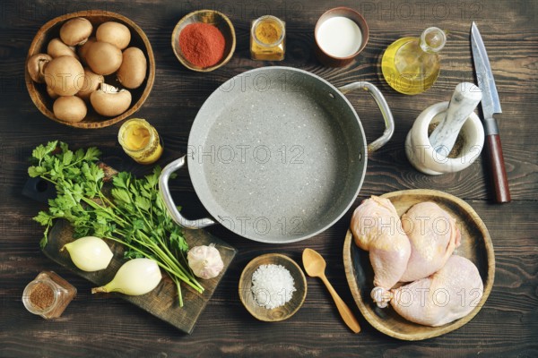 Top view of food ingredients for cooking including chicken thighs, vegetables, spices and empty pot on dark rustic wooden background