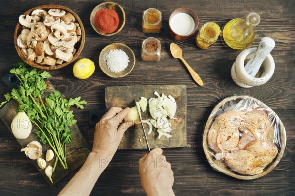 High angle view of hands slicing fresh white onion on a wooden cutting board. Chopped mushrooms, marinated chicken meat and other ingredients are on the wooden tabletop nearby