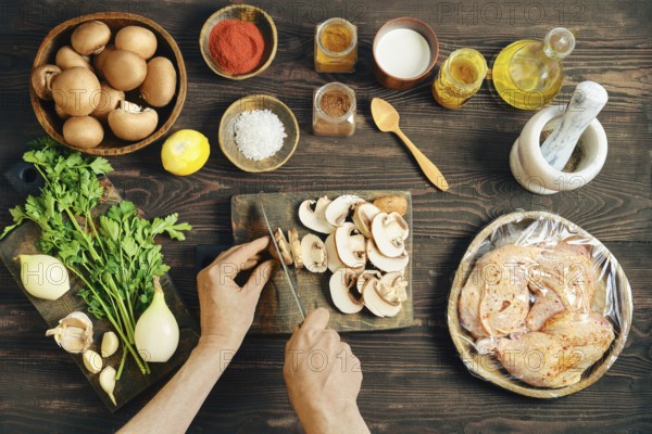 High angle view of person cutting mushroom for a meal. Lifestyle food preparation with wrapped chicken, onion, and herb on a dark rustic kitchen table