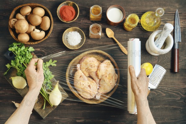 Hands wrapping marinated chicken with plastic stretch film for marinating. High angle view of rustic wooden tabletop with ingredients