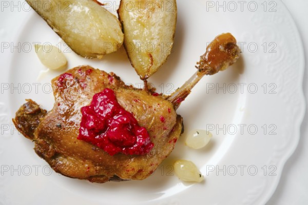Close-up view of seared duck leg on a white plate alongside roasted pears and small pearl onions. A tasty lingonberry sauce is drizzled over the duck, making the dish appealing at a dining table