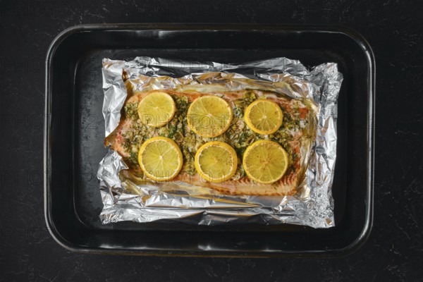 Pink salmon is placed on a baking sheet lined with foil. Lemon slices are arranged on top of the fish. The kitchen setting shows the preparation for a meal