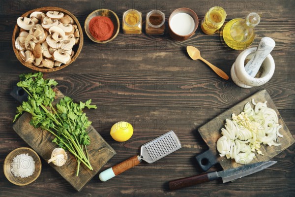 Fresh ingredients and tools for cooking are arranged on a wooden tabletop. Flat lay with chopped mushrooms, onions, herbs, spices, and kitchen tools for meal preparation