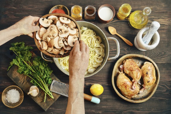 Two hands hold a bowl of sliced mushrooms over a pot with cooking onions on a wooden table. Ingredients like chicken, garlic, and herbs are arranged around the cooking area