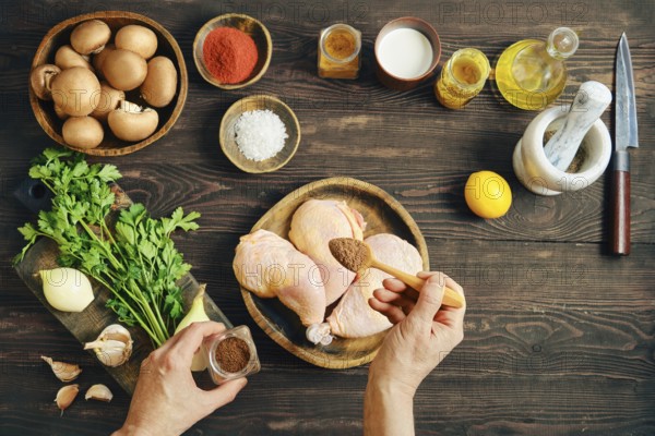 Top view of hands seasoning raw chicken legs with spicy pepper mix while preparing food on dark rustic wooden table surrounded by mushrooms, vegetables and ingredients