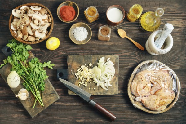 Top view of lifestyle food prep with sliced mushroom and wrapped chicken on a dark rustic wooden table. Chopped white onion and garlic are in the center od tabletop
