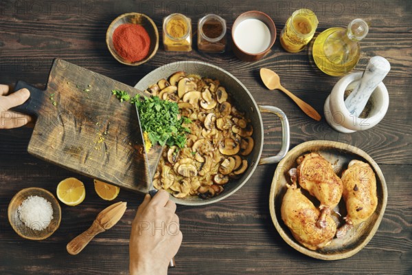 Unrecognizable person adds chopped herbs to a frying pan with fried sliced mushrooms. Nearby, bowls of spices, lemon, and roasted chicken are visible. Process of making creamy mushroom sauce
