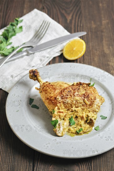 A chicken leg is placed on a gray plate. It is golden brown and garnished with green herbs. A piece of lemon is sliced and positioned nearby. Silver fork and knife are set on a napkin