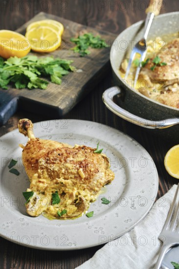 A chicken portion on a plate, surrounded by lemon slices and herbs. Cooking pan with more chicken sits nearby. Fresh parsley and lemon halves are on the wooden surface