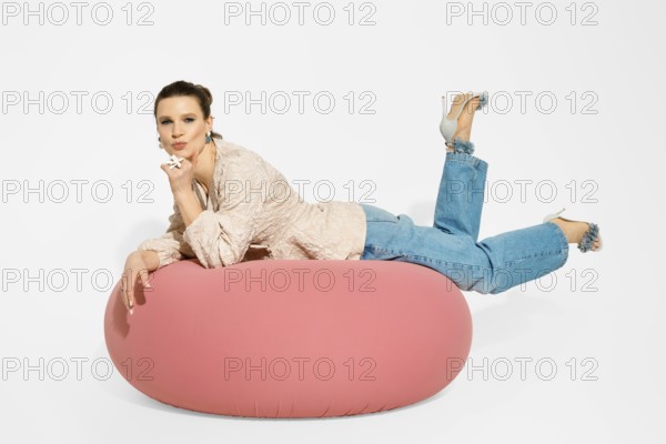 A model lies on a pink bean bag in a studio. She is wearing casual clothes with flared jeans and high heels
