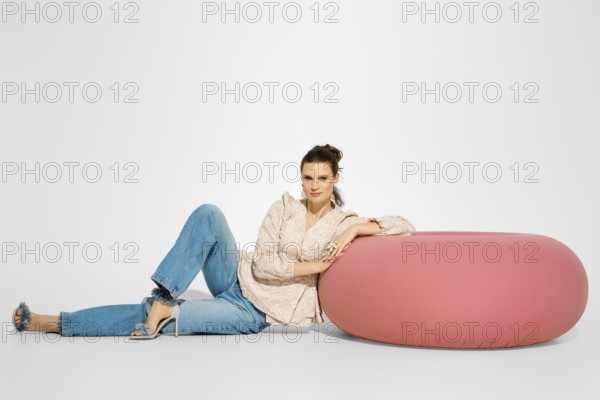 A woman in a light top and blue jeans is sitting next to a large pink cushion. She rests her arm on the cushion while posing for the camera. The setting has a plain background