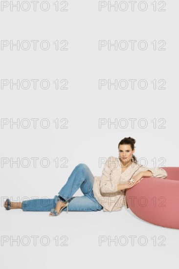 A woman sits on a floor near a pink piece of furniture in a bright studio. She leans back, wearing jeans and a light blazer. The setting is simple with a plain background and natural light coming through