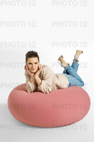 A woman lays on a big pink cushion in a studio. She is wearing a light blazer and jeans. Her hair is styled neatly. She poses with her hands on her cheeks, looking at the camera with a smile