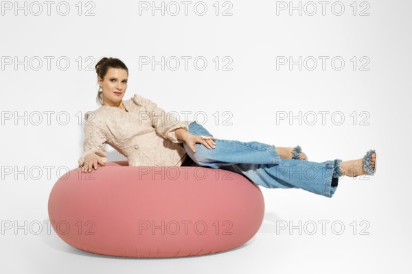 Woman is lying on a large pink lounge chair with casual clothes. The setting is a studio with bright lighting. The model displays the latest fashion trends and styles in relaxed poses