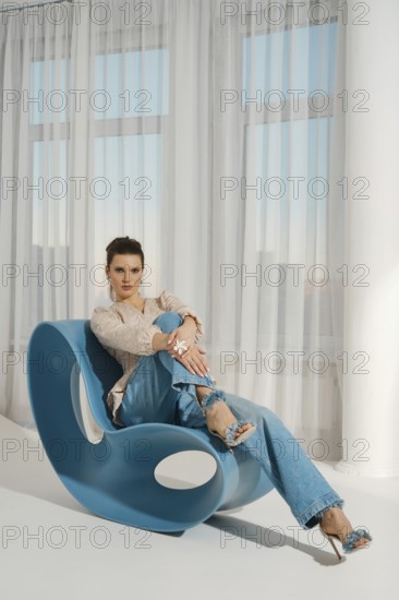 A woman is sitting on a blue chair. She is wearing casual clothes and is smiling. The room has a lot of natural light coming in through the windows. She looks relaxed and confident