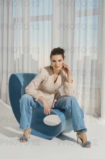 A woman sits on a blue chair with a unique design. She wears a light-colored top and blue jeans. The room has large windows that let in sunlight, creating a warm atmosphere