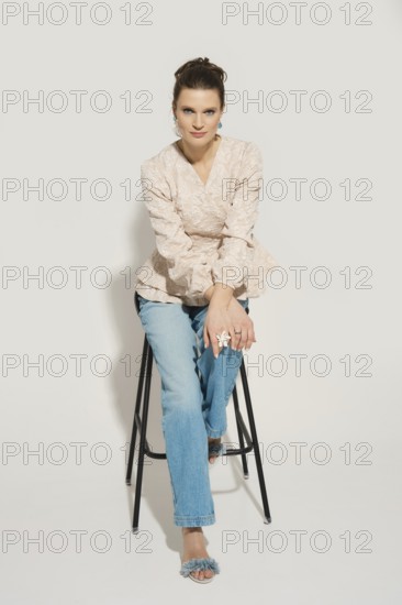 A model sits on a tall stool in a studio. She wears casual clothes including a light top and denim jeans. The background is plain and highlights her outfit as she poses for the photoshoot