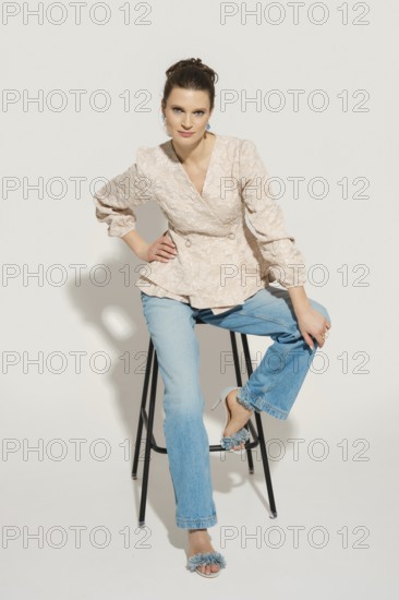 A model poses on a tall stool in a studio with a plain background. She wears a fashionable top and jeans paired with peep-toe heels. The setting is well lit, highlighting the outfit and her pose