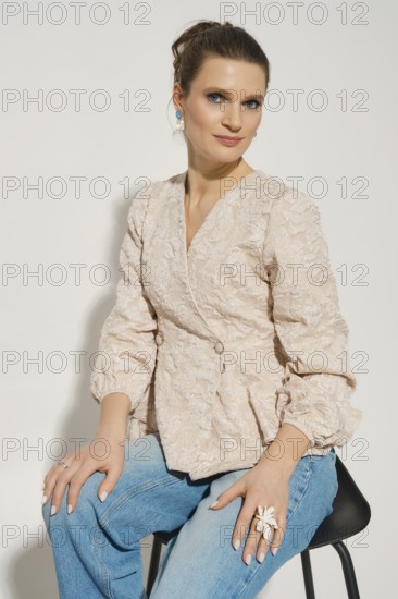 A woman sits on a black chair wearing a light-colored blazer and blue jeans. She has her hair styled and poses with a relaxed expression. The background is simple and neutral, focusing on her attire
