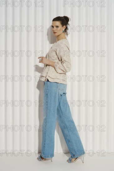 A model stands sideways with her left foot forward, wearing a casual outfit consisting of a light top and blue jeans. She is posed in front of a white striped backdrop