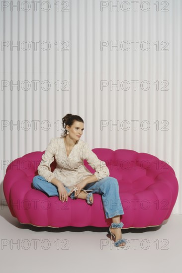 A woman is sitting on a large pink sofa in a modern indoor area. She wears a light-colored blazer and jeans. The setting features a simple striped wall and natural light coming from above