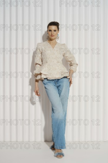 Relaxed woman stands with one hand on her hip. She wears a light blouse and blue jeans. The background has vertical lines creating a clean studio look. Natural light illuminates the scene