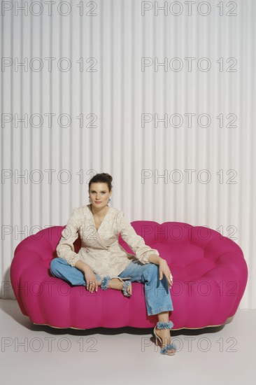 Woman sits with one leg tucked under her on a pink couch in a studio. She wears a light blazer and blue jeans. The background features vertical lines, creating a modern design