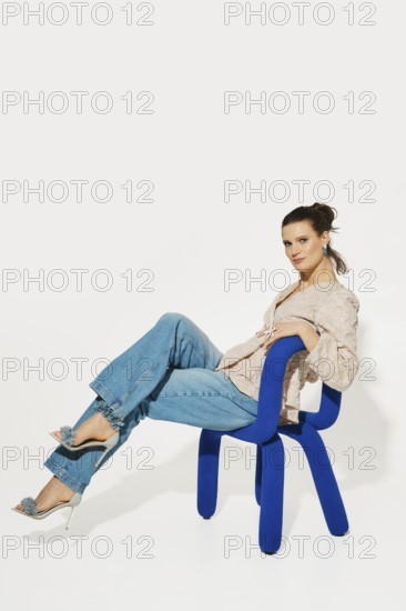 A model sits on a blue chair, leaning back with one leg crossed over the other. She wears a light shirt and loose jeans. The background is plain and bright. The focus is on her pose and outfit