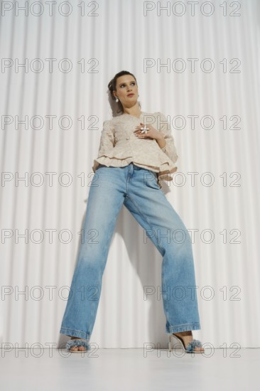 A woman stands confidently in a relaxed pose wearing wide-leg jeans and a light blouse. She is in a modern indoor space with a textured white wall. Her styling is simple, showcasing her outfit