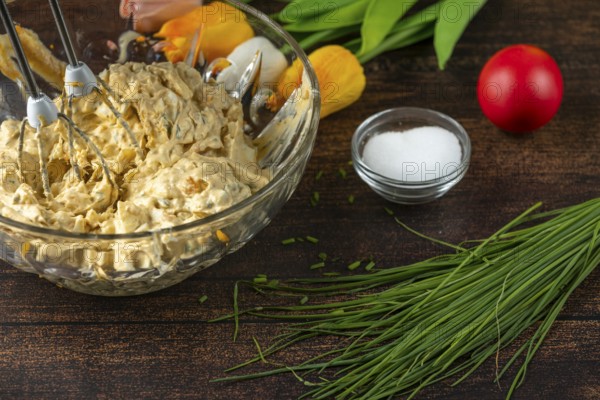 Egg salad in a glass bowl with a mixer, fresh ingredients in the background