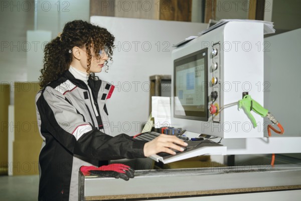 A woman is using a computer in a warehouse. She wears work gear and looks focused while interacting with the screen. Items are nearby, showing a busy work environment