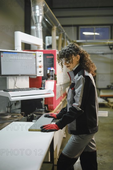 Woman wearing protective gloves and safety glasses runs a wooden piece into a machine for gluing with edge tape. The factory has machinery and tools visible in the background