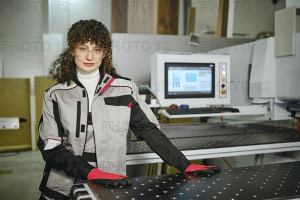 Portrait of female worker next to a large cutting machine in a furniture workshop. She wears gloves and safety glasses