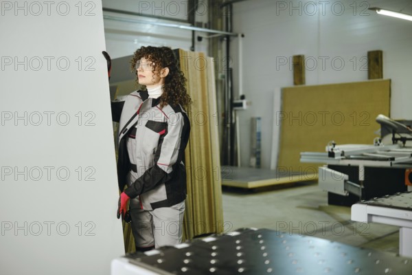 A worker prepares to cut a large sheet of wood in a workshop. The worker is dressed in protective work gear and uses tools for the task. Various materials and machines are present in the background