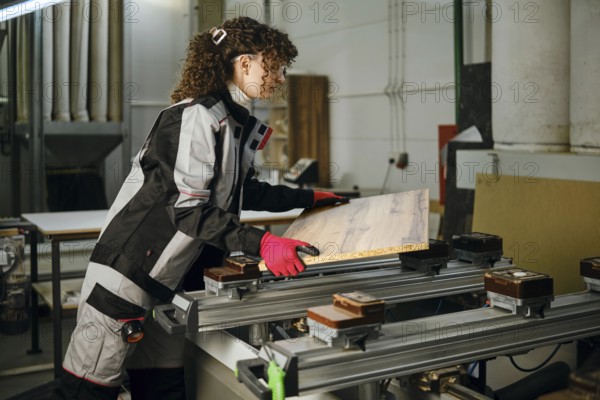 A woman is in carpentry workshop handling a piece of wood. She wears safety gloves and works with machinery. Tools and equipment are placed around her. The scene shows activity and focus on the task
