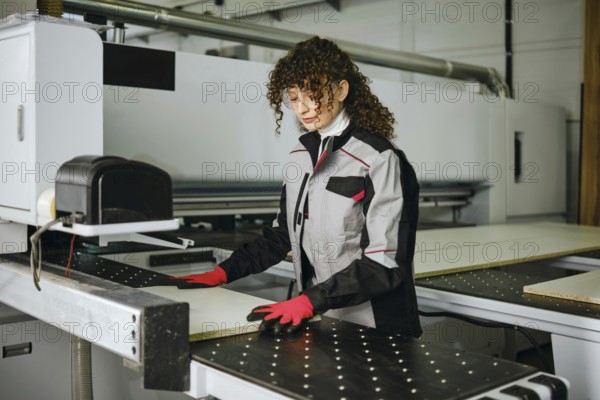 A woman stands at a CNC machine in a factory. She puts a wooden sheet in a sawing machine. She is focused on her task while wearing gloves and safety glasses