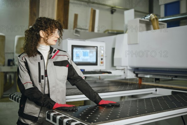 A woman stands beside wood cutting machine in carpentry workshop. She wears gloves and protective glasses while working with wood cutting machine