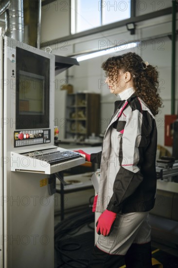 Woman stands at a control panel in a manufacturing facility. She wears a work uniform with gloves. The machine is active, showing data on the screen