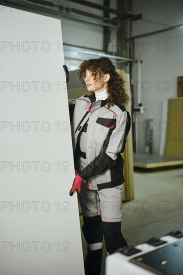 A woman is positioning a large wooden panel carefully in carpentry workshop. The area is filled with tools and materials