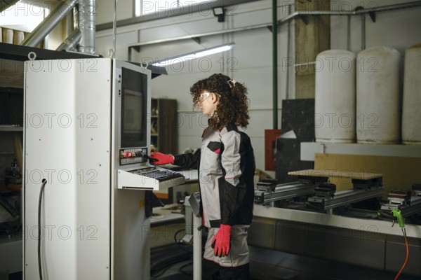 A person operates a milling machine in a carpentry workshop filled with tools and equipment. The worker wears gloves and stands by the control panel, focused on the task
