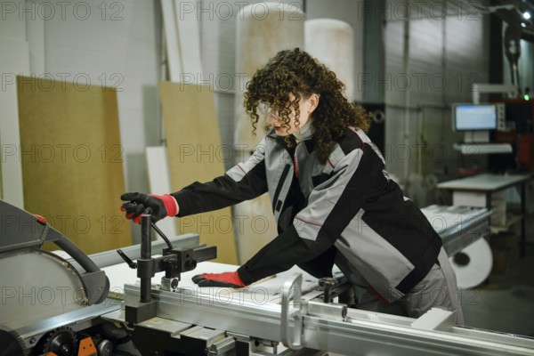 A woman in a workshop operates a table saw machine. She uses her hands to adjust a piece of wood. The setting features various tools and materials, showing a busy manufacturing environment