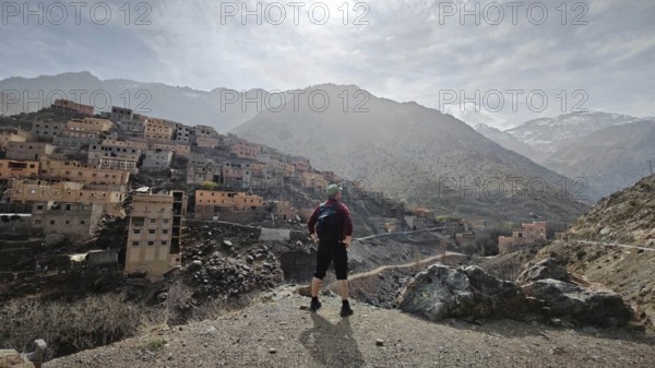 A hiker with backpack stands in front of a mountainous village in sunny weather, Atlas Mountains, Morocco