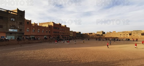 Children playing soccer on a sandy playing field between buildings in cloudy sky, Morocco