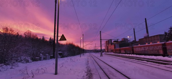 Sunrise at a snow-covered train station on Rennsteig, railroad tracks in purple sky with factory in the background, Frankenwald nature park Park