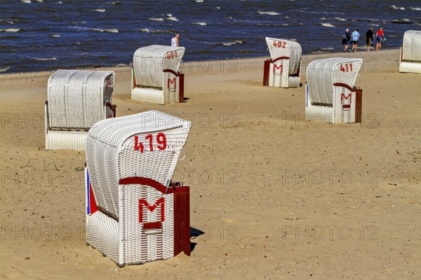 Several white beach chairs with red accents stand on the beach with walkers in the background, beach chairs on the beach in Cuxhaven