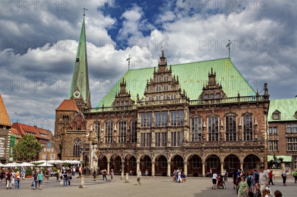 Large Renaissance building next to church tower, surrounded by people in a busy square under cloudy sky, The Town Hall of Bremen