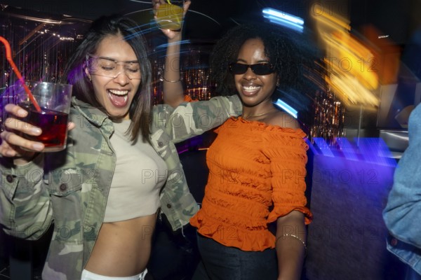 Diverse young women are having fun, raising drinks, and dancing together in a lively nightclub setting with vibrant neon lights and motion blur, enjoying their night out