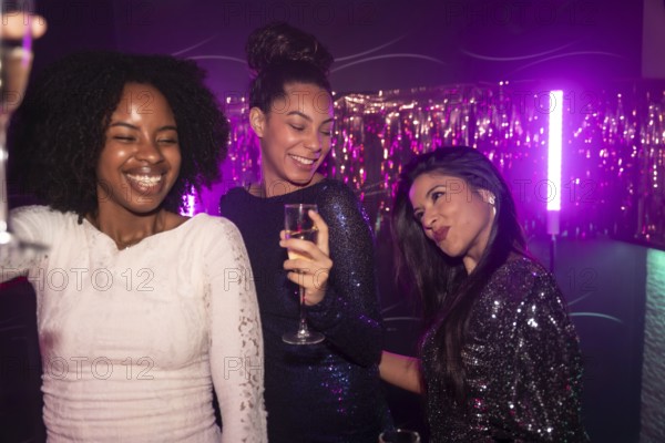 Three diverse women friends enjoying a night out, toasting with champagne and smiling while celebrating in a vibrant nightclub under purple neon lights with tinsel decorations